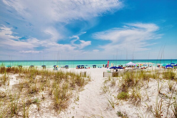 Ein Strand mit einem sandigen Pfad zum Wasser, Sonnenschirmen und Liegestühlen.