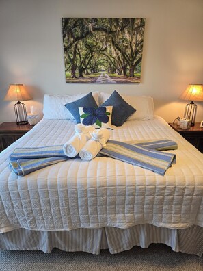 A neatly made bed with a striped quilt, pillows, and towels, a painting of trees on the wall, and two bedside lamps. - Peaceful Lakefront Retreat | 5* View, Dock, Games (Eatonton)