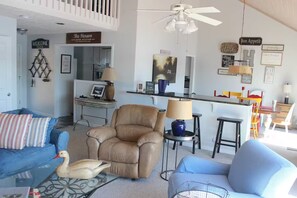 A living room with a brown leather chair, a blue sofa, a glass coffee table, and a swan figurine. There is a kitchen area in the background with a sign that reads 'The Henson Lake House.' - Peaceful Lakefront Retreat | 5* View, Dock, Games (Eatonton)
