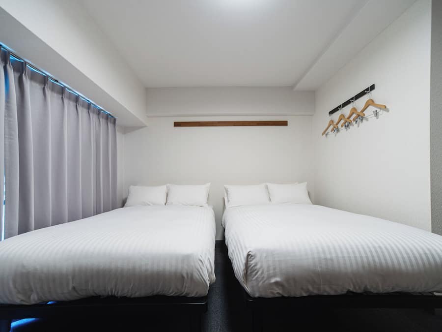 Um quarto com duas camas, uma janela com cortinas, uma prateleira na parede e um cabideiro.