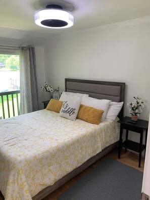 A bedroom with a bed, a nightstand, a potted plant, and a view of the outdoors.
