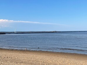 A sandy beach with calm water, a distant sailboat, and a rocky breakwater. - Sea Esta by the Marsh: 2 minutes to the beach! (Biddeford)