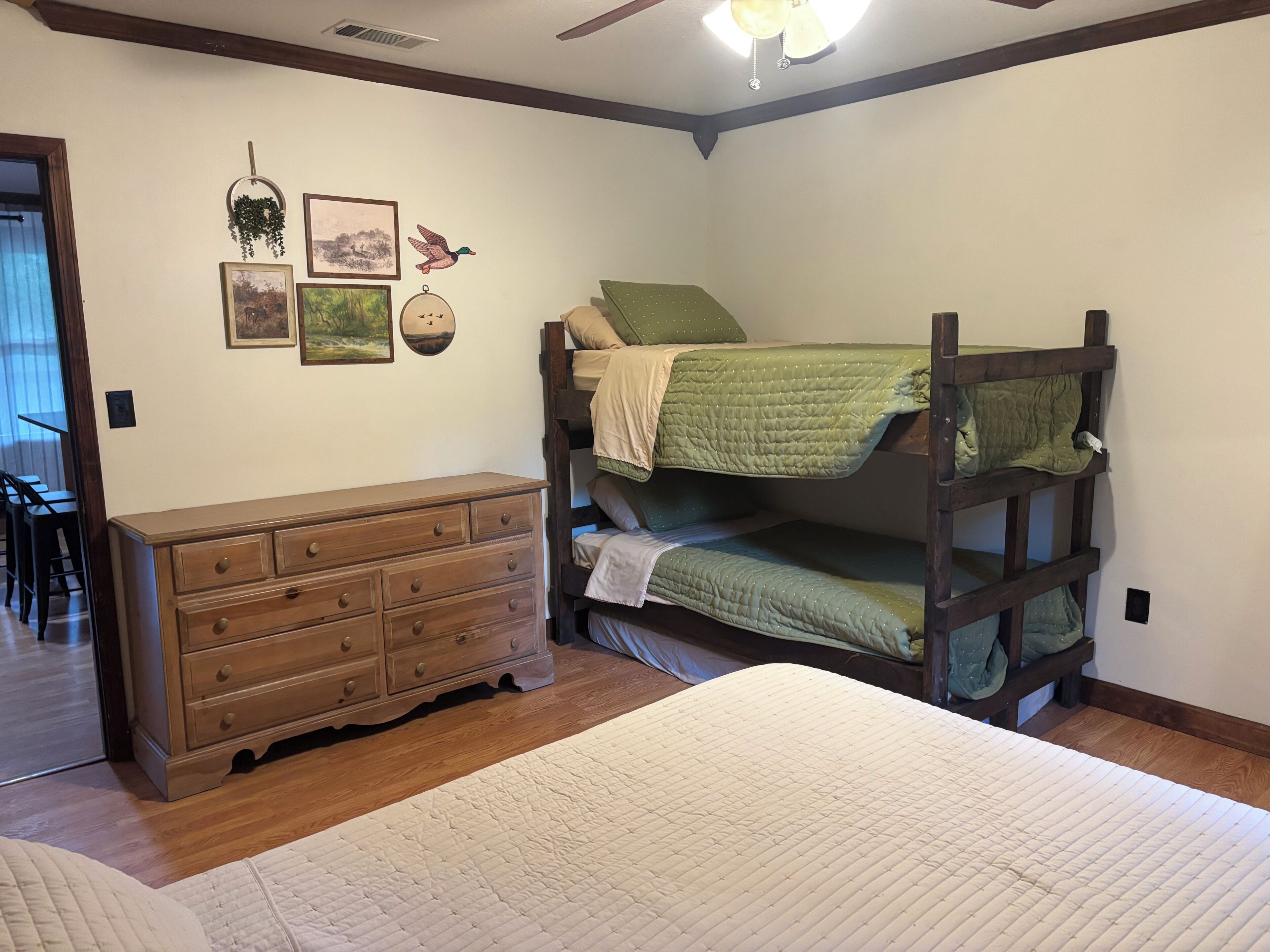 Un lit superposé avec une couverture verte, une commode en bois et des décorations murales comprenant un canard et des tableaux encadrés.