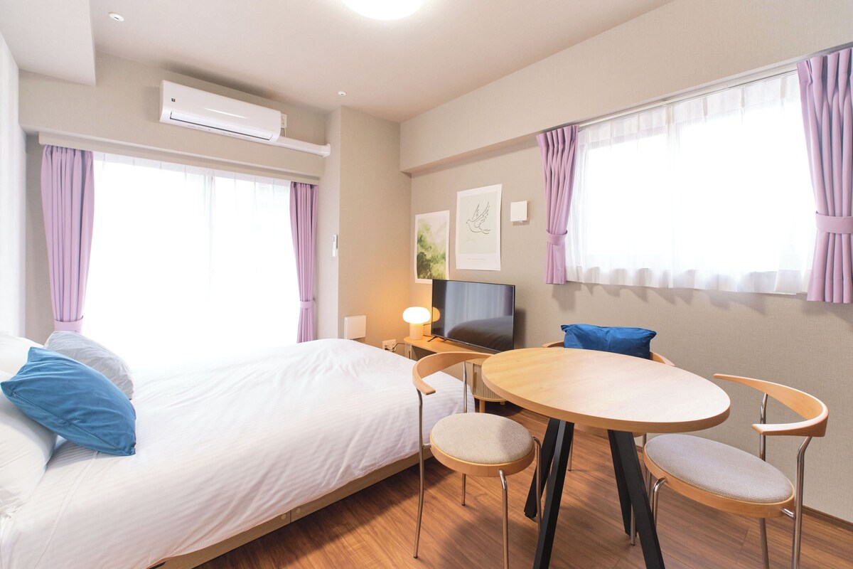 Une chambre d’hôtel comprenant un lit, un bureau avec une télévision, une table ronde avec des chaises et de grandes fenêtres avec des rideaux.