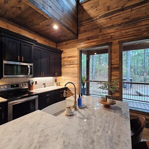 A kitchen with dark cabinets, a stainless steel microwave, a sink, and a large window overlooking a forested area. - Luxe Cabin, 4BR, Hot Tub, ATV Trail, Dog Friendly (Broken Bow)
