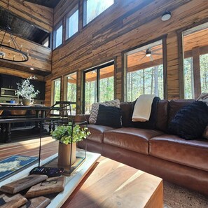A cozy cabin interior with a wooden ceiling, a leather sofa, a dining area, and large windows. - Luxe Cabin, 4BR, Hot Tub, ATV Trail, Dog Friendly (Broken Bow)