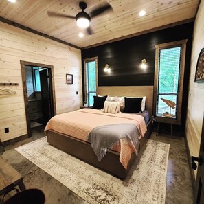 A bedroom with a bed, a ceiling fan, a window with blinds, and a door leading to another room. - Luxe Cabin, 4BR, Hot Tub, ATV Trail, Dog Friendly (Broken Bow)