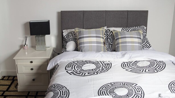 Een net opgemaakt bed met een bedsprei met een geometrisch patroon, een witte nachtkastje met lades, een lamp en een decoratieve doos.