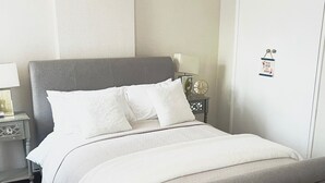 Una cama bien hecha con un cabecero gris, una mesita de noche con una lámpara y un reloj de pared.