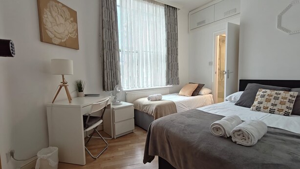 A hotel room with a desk, two beds, a window with curtains, a radiator, and a door. - Eurostar Private Flat Sleeps up to 10 London (London)