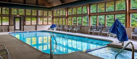 An indoor swimming pool with a wooden ceiling, clear windows, and lounge chairs.