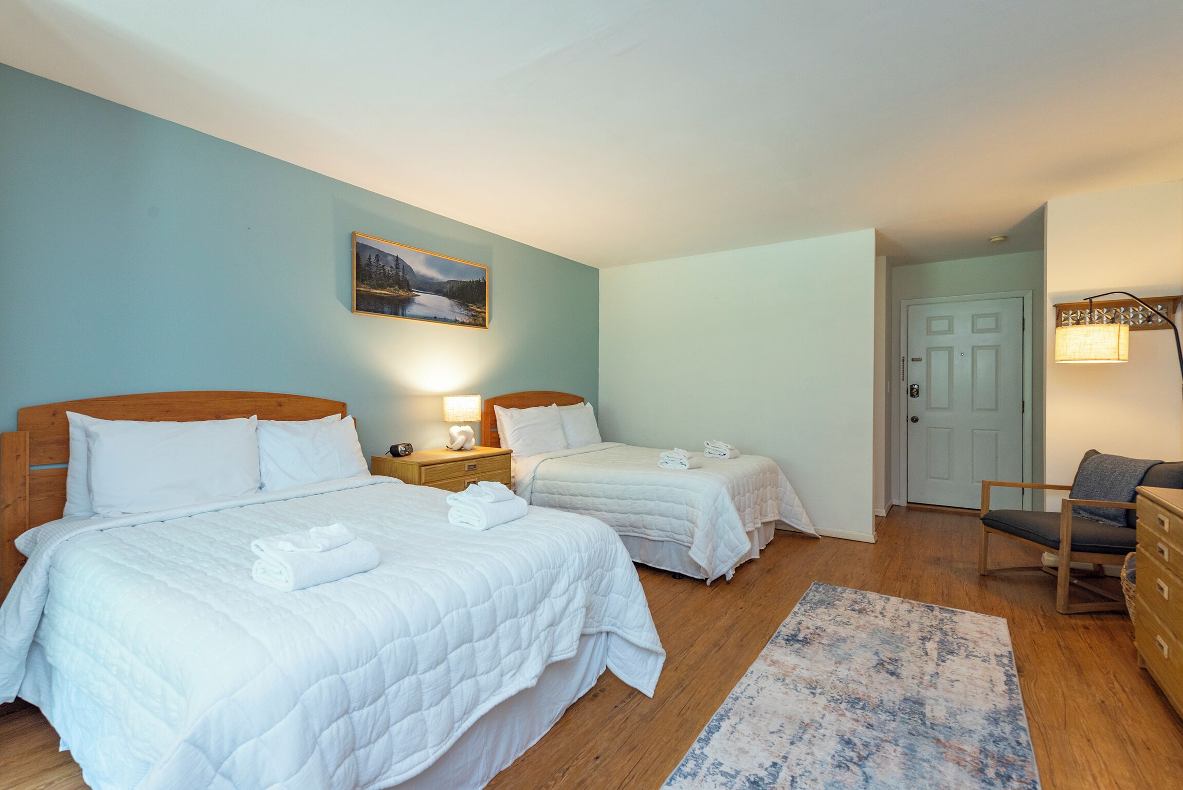 A hotel room with two beds, a nightstand, a lamp, a painting, a chair, and a door.