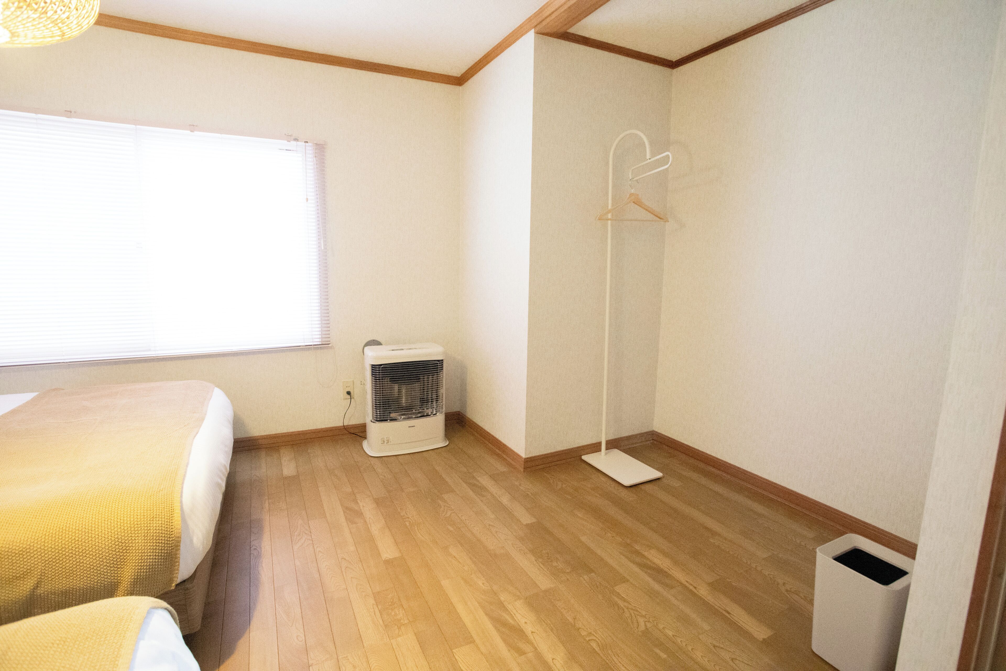 A room with a bed, a standing heater, a floor lamp, and a trash bin.