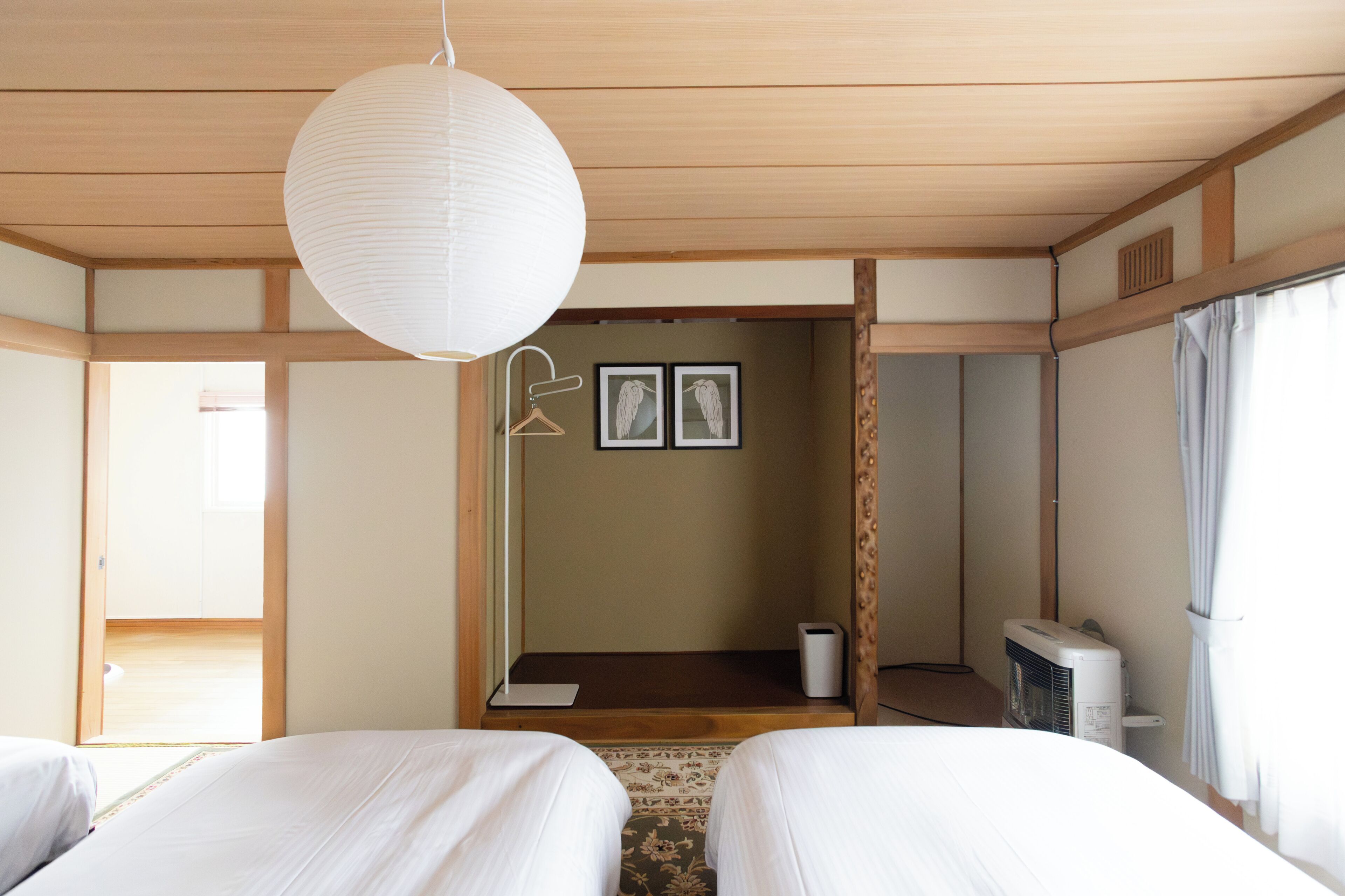 A hotel room with two beds, a hanging paper lantern, a small desk, a wall-mounted heater, and a view of the outside through a window.