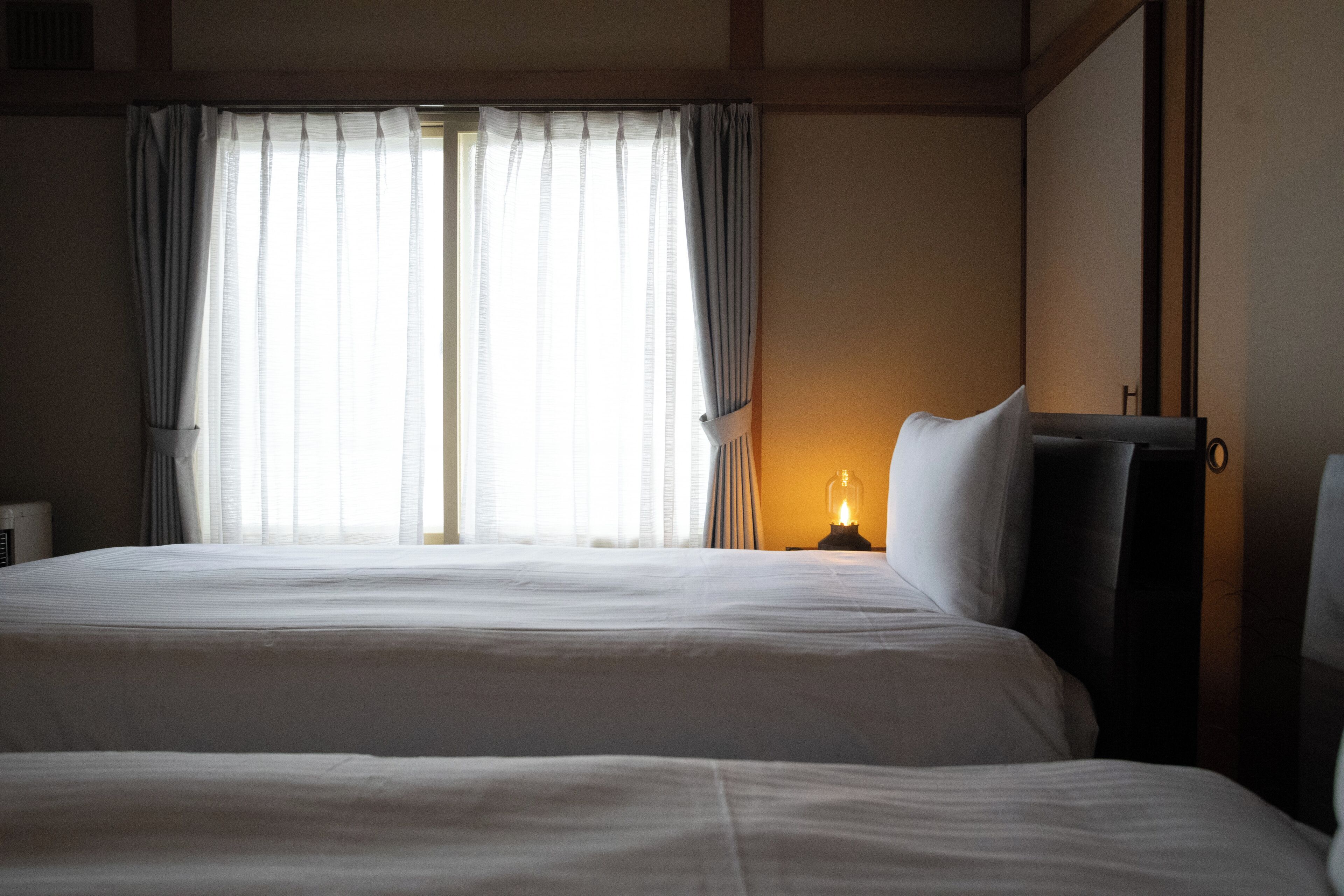 A hotel room with two beds, a nightstand with a lamp, and a window with curtains.