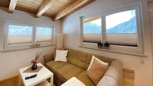 Una habitación con techo de madera, dos ventanas con persianas, un sofá, una mesita con un florero y un control remoto.