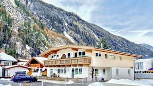 Un hotel de varios pisos con balcón, un automóvil estacionado adelante y una montaña nevada al fondo.