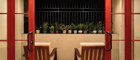 A room with two wooden chairs, a window with barred security, and a row of potted plants.