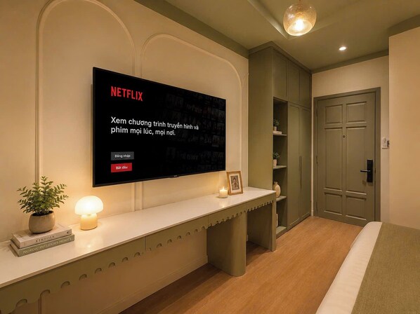 Una habitació d'hotel moderna amb un gran televisor de pantalla plana penjat a la paret, una taula de nit amb una làmpada, una fotografia emmarcada i una planta en test.
