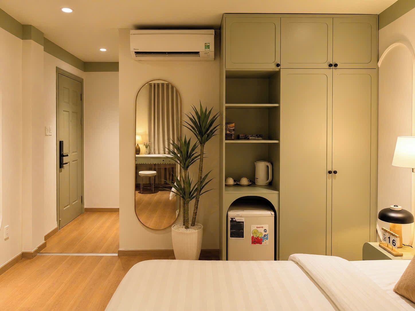 Habitación de hotel con cama, escritorio con silla, espejo, planta, refrigerador pequeño y armario.
