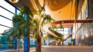 A reception area with a curved reception desk, palm trees, and a staircase leading to an upper level. - Baltic Zinnowitz (Zinnowitz)