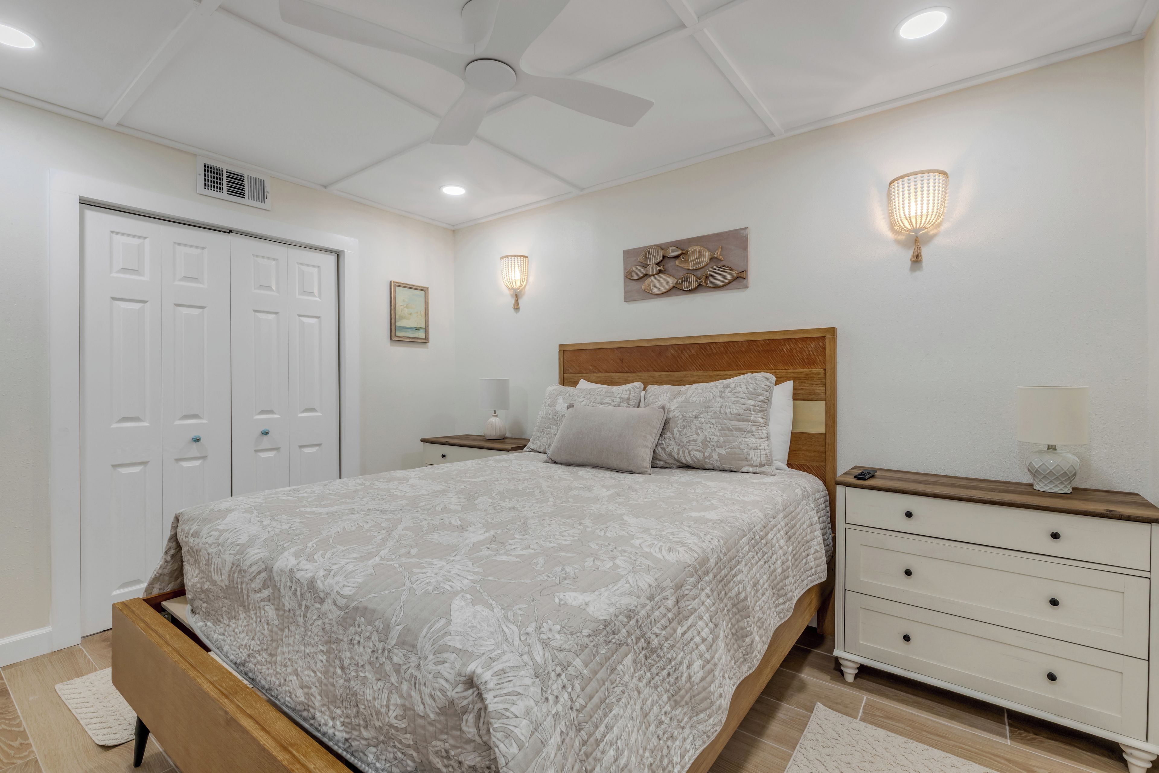 Ein Schlafzimmer mit einem Bett, Nachttischen, einem hölzernen Kopfteil, einem Deckenventilator, Wandleuchten und einer Tür.