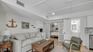 A modern living room with a sofa, coffee table, and armchair, a kitchen area with cabinets, and a dining table. - Condo 12 Coastal Comfort (New Smyrna)