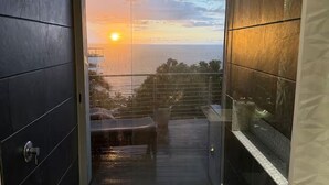 Parveke, jolta avautuu näkymä auringonlaskuun meren yllä, lasiovi ja pieni oleskelualue.