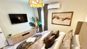 A modern bedroom with a flat-screen TV, a bed with multiple pillows, a bedside table with a plant, and an air conditioning unit. - LUXURY DOWNTOWN COLUMBUS TOWNHOME (Columbus)