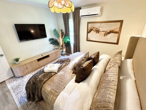 Ein modernes Schlafzimmer mit Flachbildfernseher, einem Bett mit vielen Kissen, einem Nachttisch mit Pflanze und einer Klimaanlage.