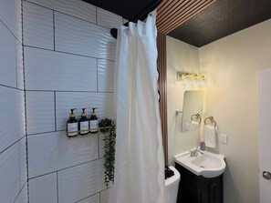 Ein Badezimmer mit weiß gefliesten Wänden, einer Duschvorhang, einem Waschbecken mit Spiegel und Pflegeprodukten auf einem Regal.