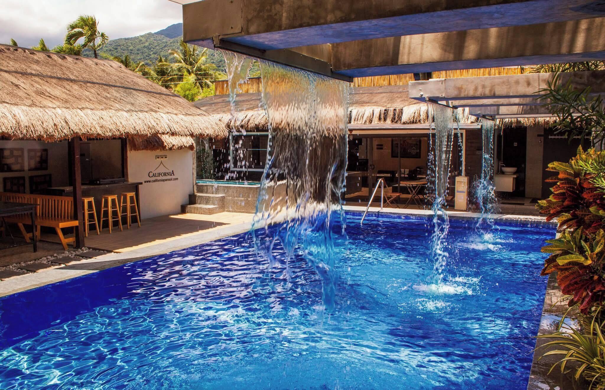 A swimming pool with water cascading from a structure above, surrounded by a thatched-roof building and wooden seating.