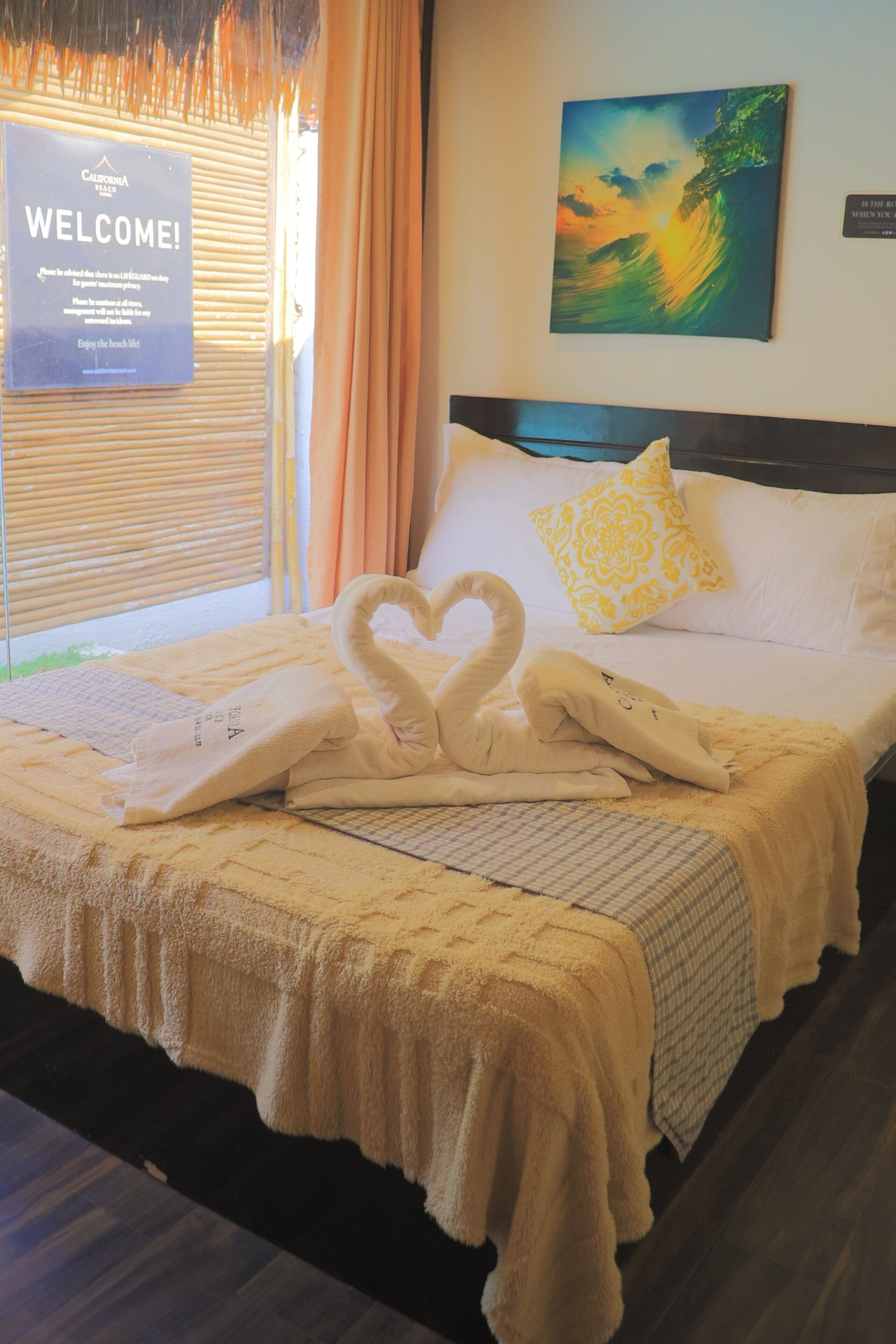 A hotel room with a bed, a welcome sign, a painting, and a swan towel arrangement.