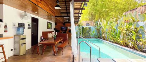 A pool area with a wooden deck, a table, chairs, a water dispenser, and a television.