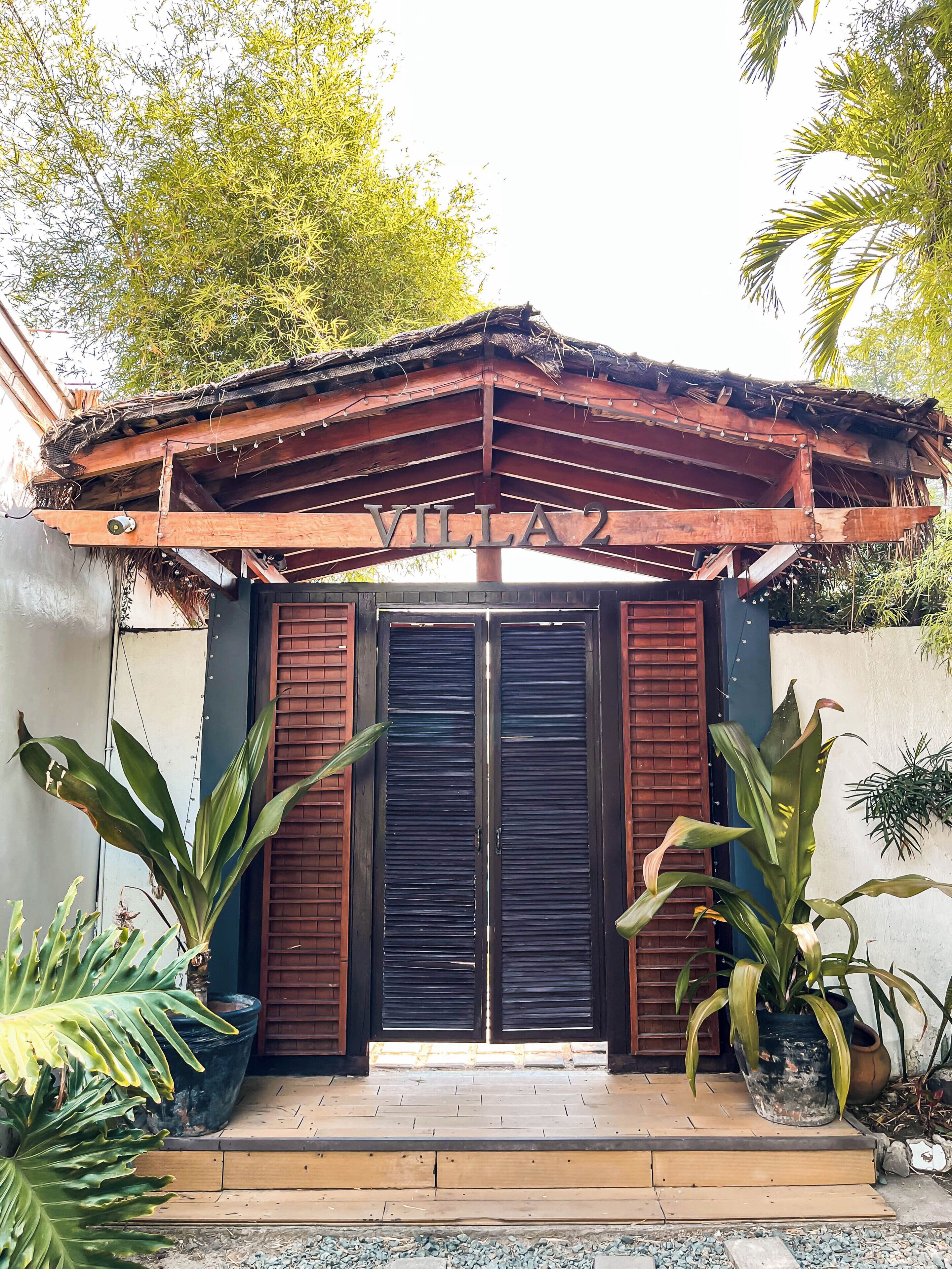 Entrada da villa com um arco de madeira, um portão preto e plantas em vasos.