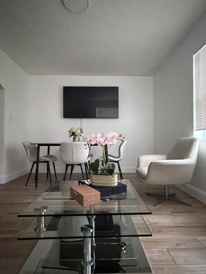 Ein modernes Wohnzimmer mit einem Glastisch, einem flachbildfernseher an der Wand und einem Blumentopf mit rosa Blüten.