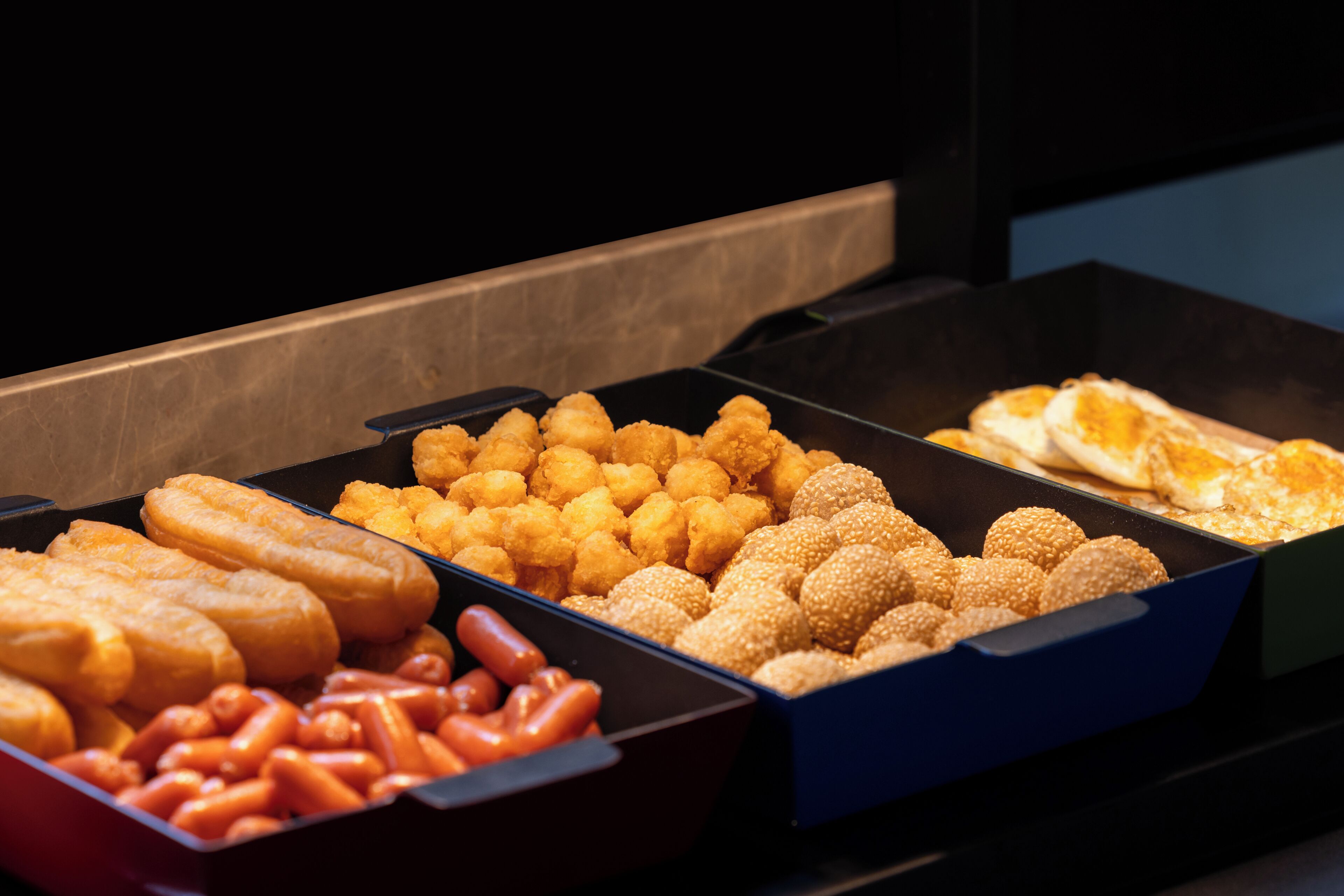 Ein reichhaltiges Buffet mit verschiedenen Speisen, darunter Würstchen, Kartoffel-Tots und Brötchen.