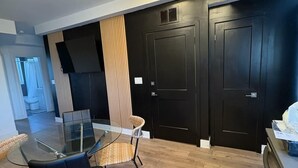 Cozinha moderna com mesa de jantar de vidro, cadeiras pretas e chão de madeira. Três portas pretas e uma parede preta com painel de madeira vertical.
