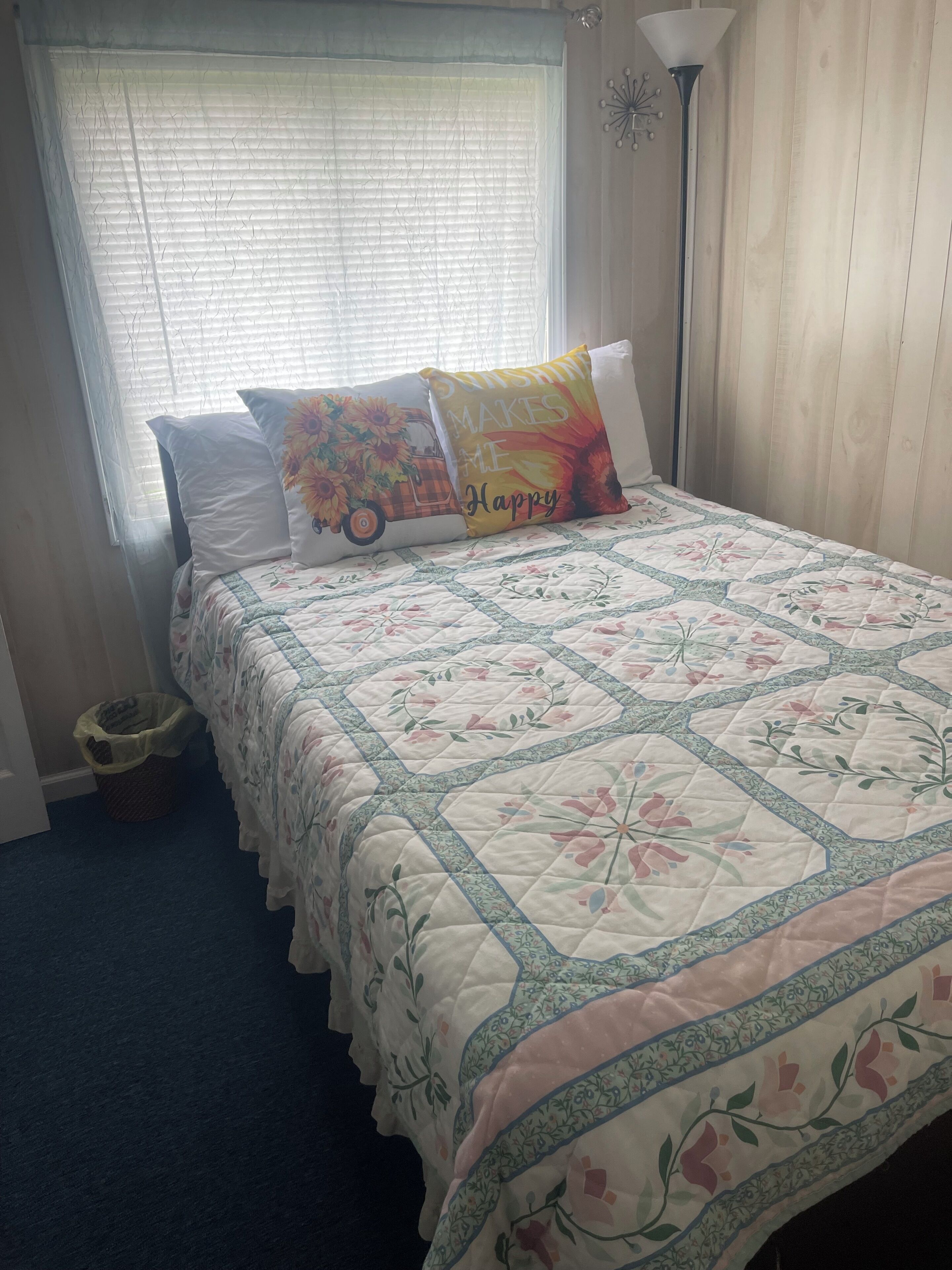 A bedroom with a bed, a window with blinds, a floor lamp, and a wall clock.
