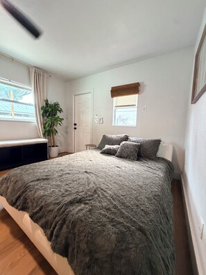 Quarto com cama, planta em vaso, janela com cortinas, porta e uma pequena mesa.
