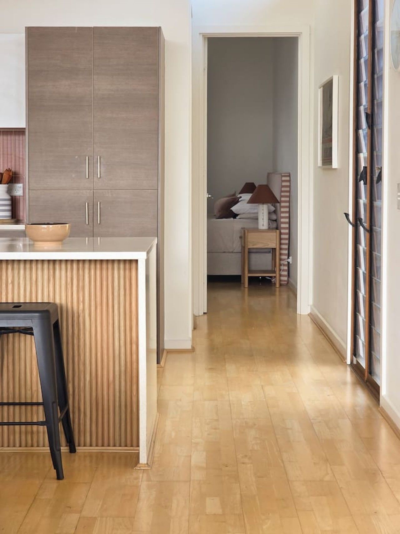 A modern kitchen with a built-in counter and cabinets, a dining area with a stool, and a hallway leading to a bedroom with a bed and bedside table.