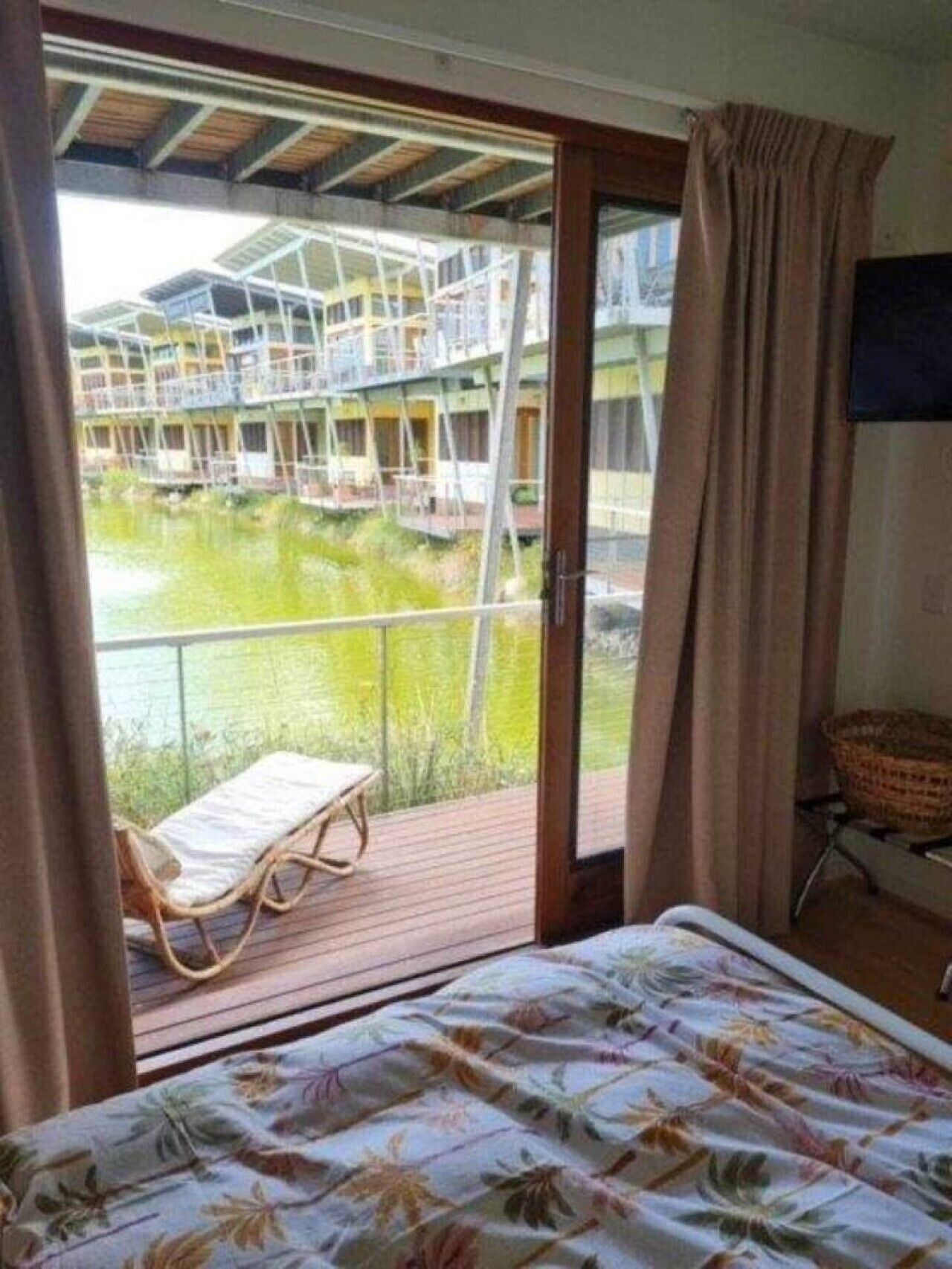 A room with a bed, a wooden deck, a view of a building with a green roof, and a window with curtains.