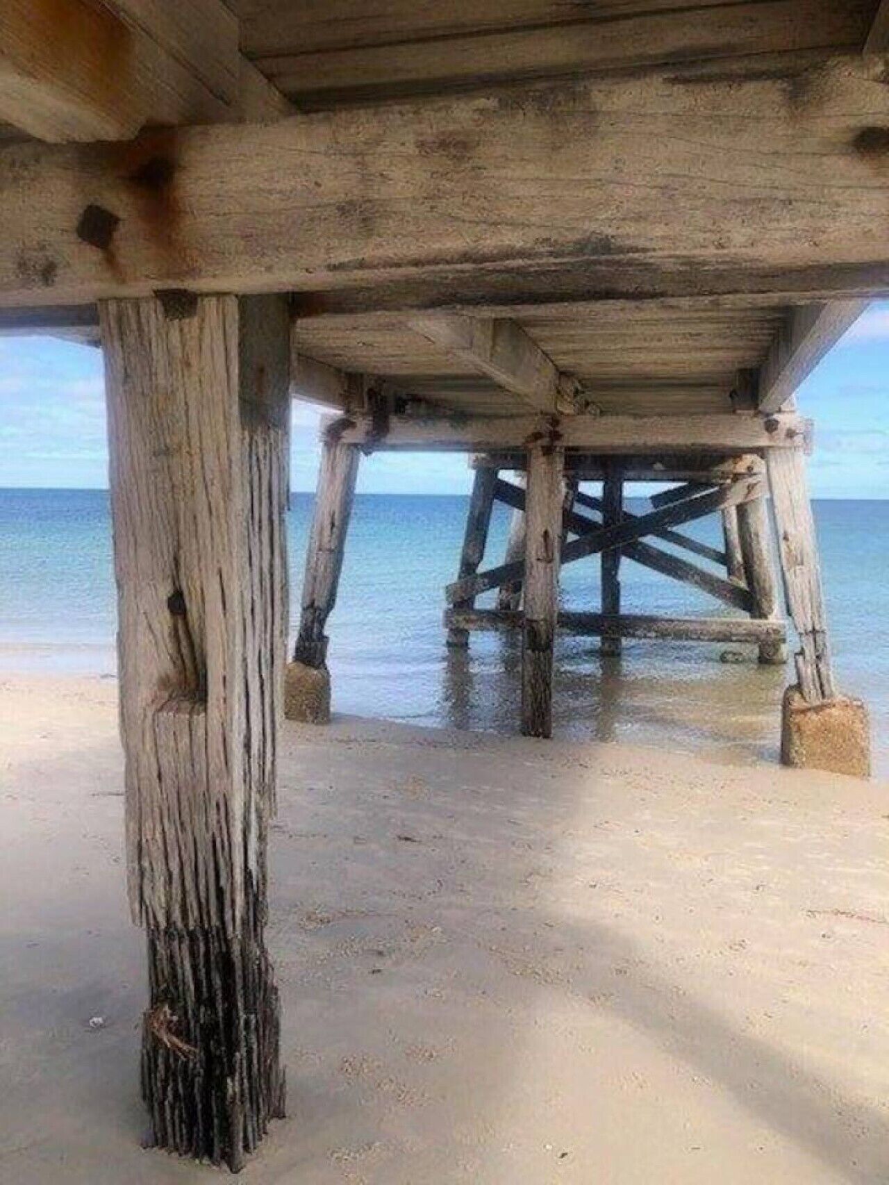 A wooden pier structure with a sandy beach and calm water in the background.