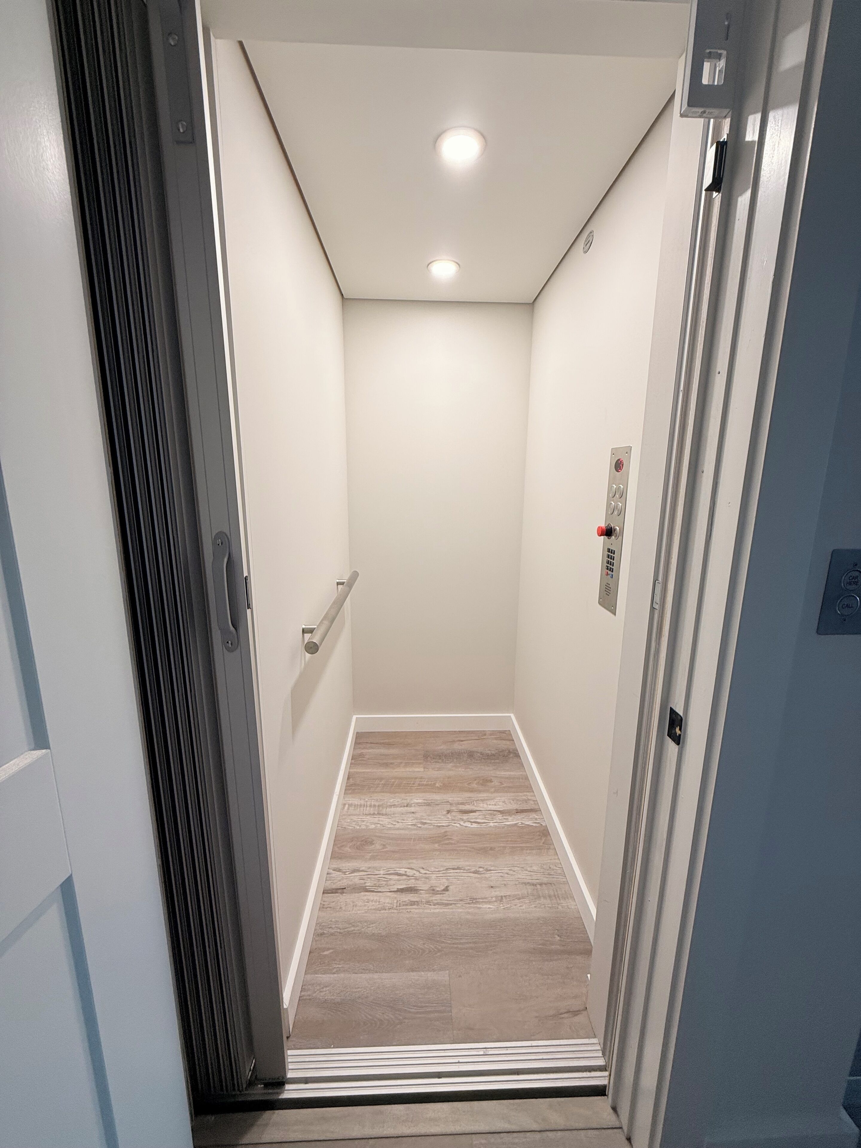 Un étroit puits d’ascenseur avec un plancher en bois, des murs blancs et un luminaire au plafond.