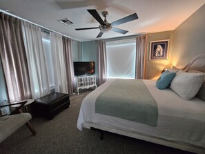 Et hotelværelse med seng, fjernsyn, ventilator og et vindue med gardiner.
