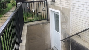 Un edificio residencial con fachada de ladrillo, rellano de concreto, barandilla metálica y puerta blanca.
