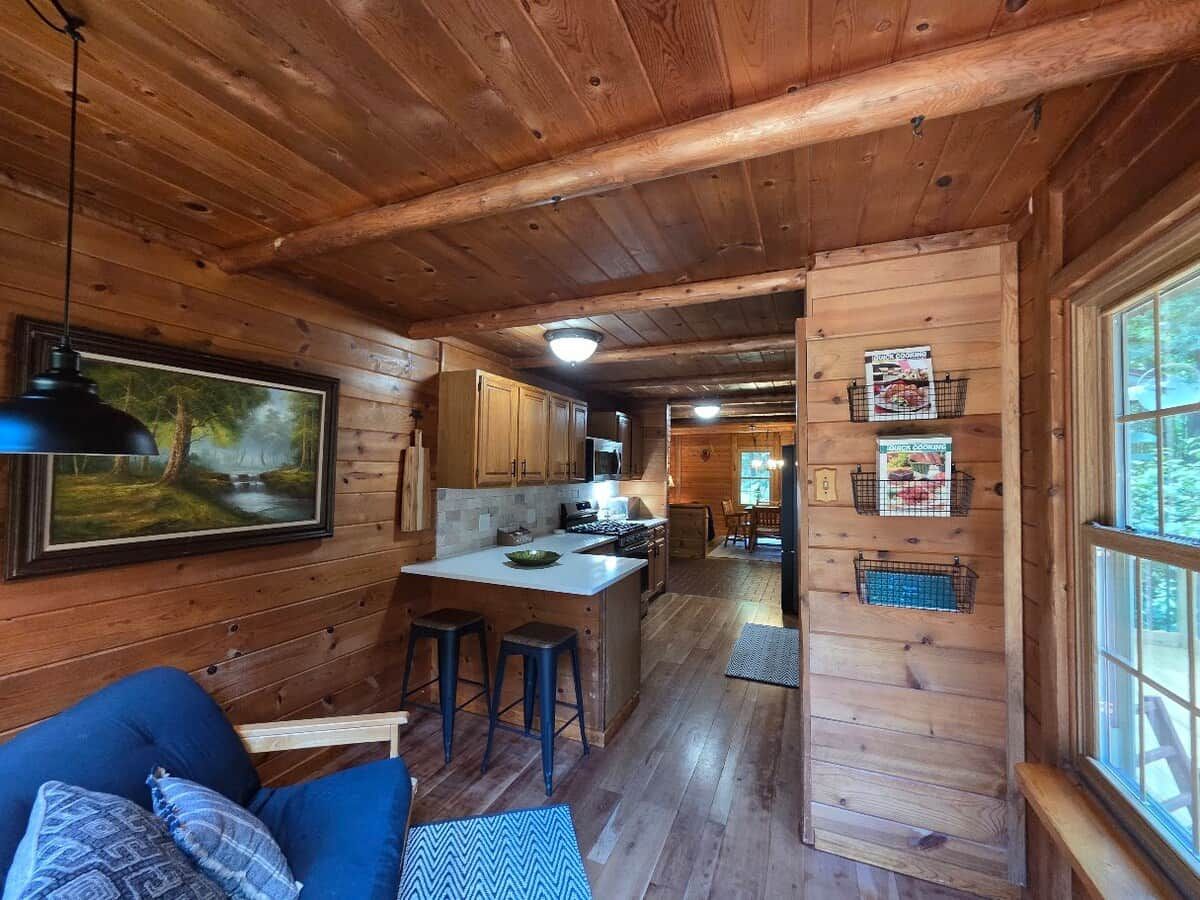 A cabin interior with a kitchen area, wooden walls, a dining area, and a large window.