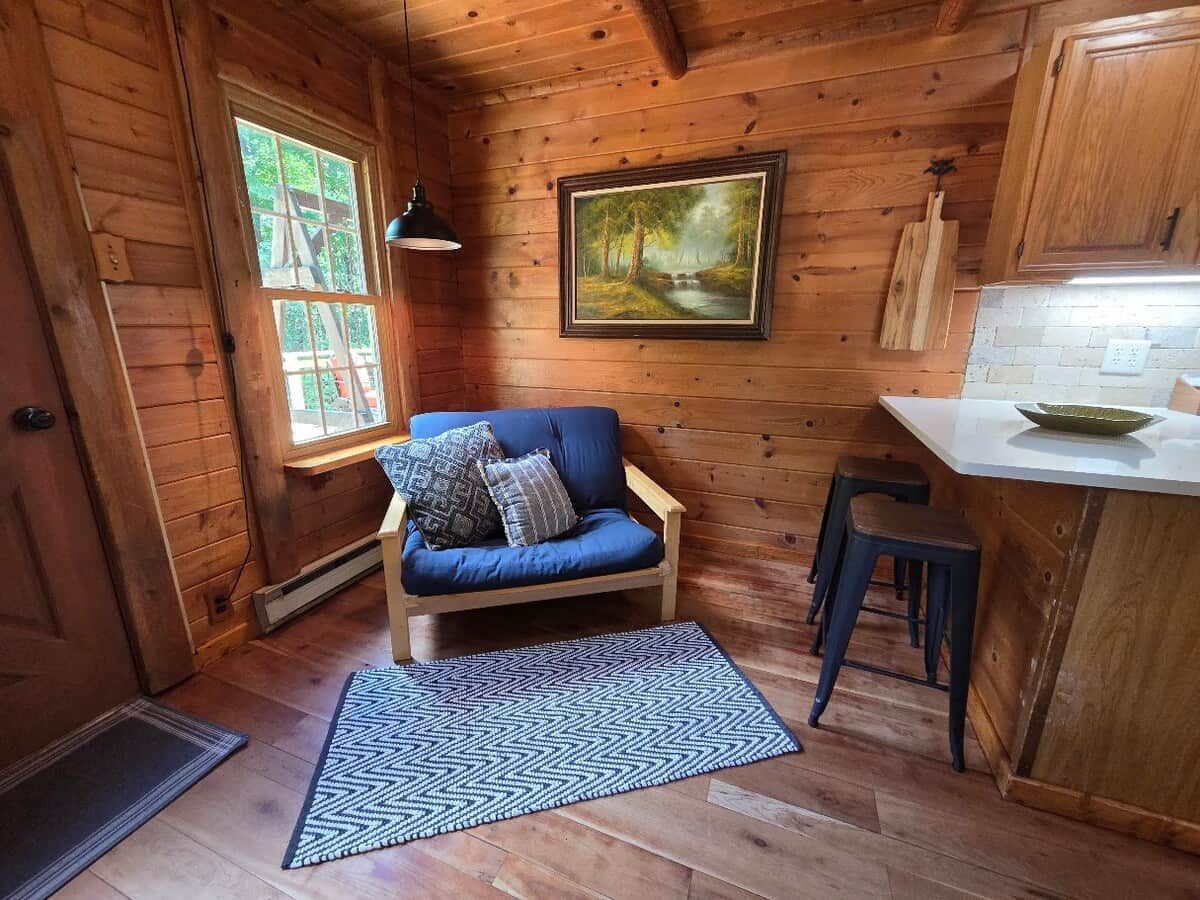 Ein Interieur einer Holzhütte mit Holzboden, einem blauen Sofa mit Kissen, einem Gemälde an der Wand und einer kleinen Küchenzeile.