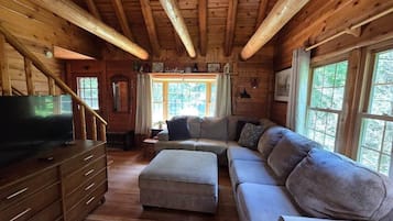 A log cabin interior with a wooden staircase, a sofa set, a television, and a window with a view of trees.
