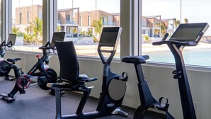 A row of stationary bikes with attached screens, positioned in a room with large windows overlooking a beach and modern buildings. - Beachfront 1 Bedroom | Outdoor Pool | Stunning Caribbean Views (Long Bay Hills)